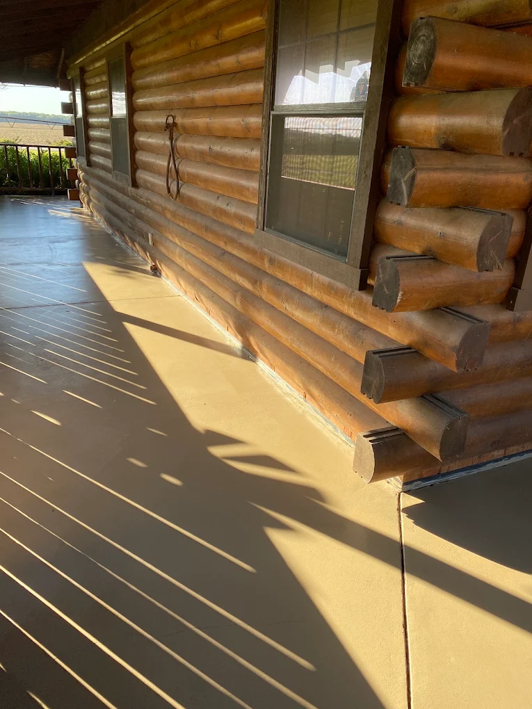 Tan coated concrete porch running along the wall of a log cabin in the afternoon sun