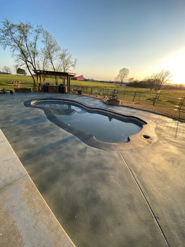 Wide, level concrete deck poured around a free-form pool with open pasture behind it at sunset